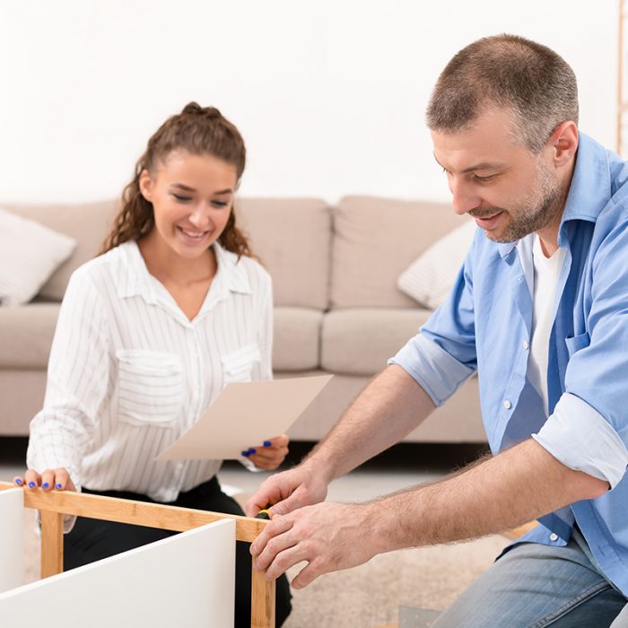 Happy Couple Installing Self-Assembly Furniture Kit Together Furnishing Home Sitting In Living Room Indoors. Handyman Service Advertisement Concept. Selective Focus