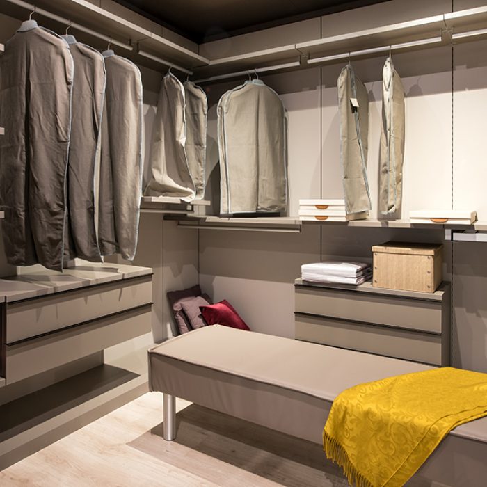 Interior of a walk in closet with hanging clothes and built in drawer units and seating in a luxury modern home