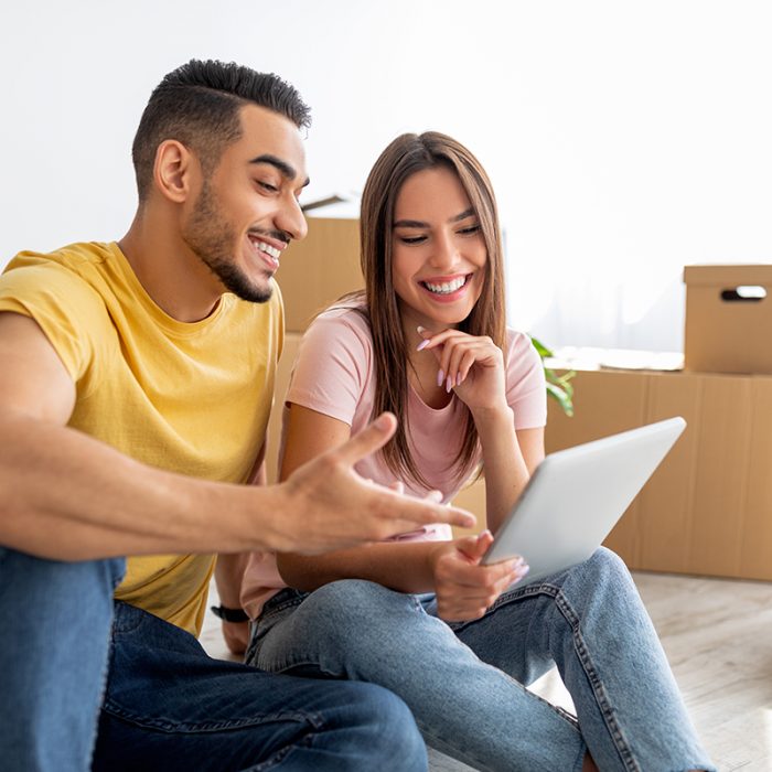Cheerful multiracial couple with tablet computer browsing web to buy household goods for new home. Cheery young tenants with touch pad sitting on floor among carton boxes on moving day